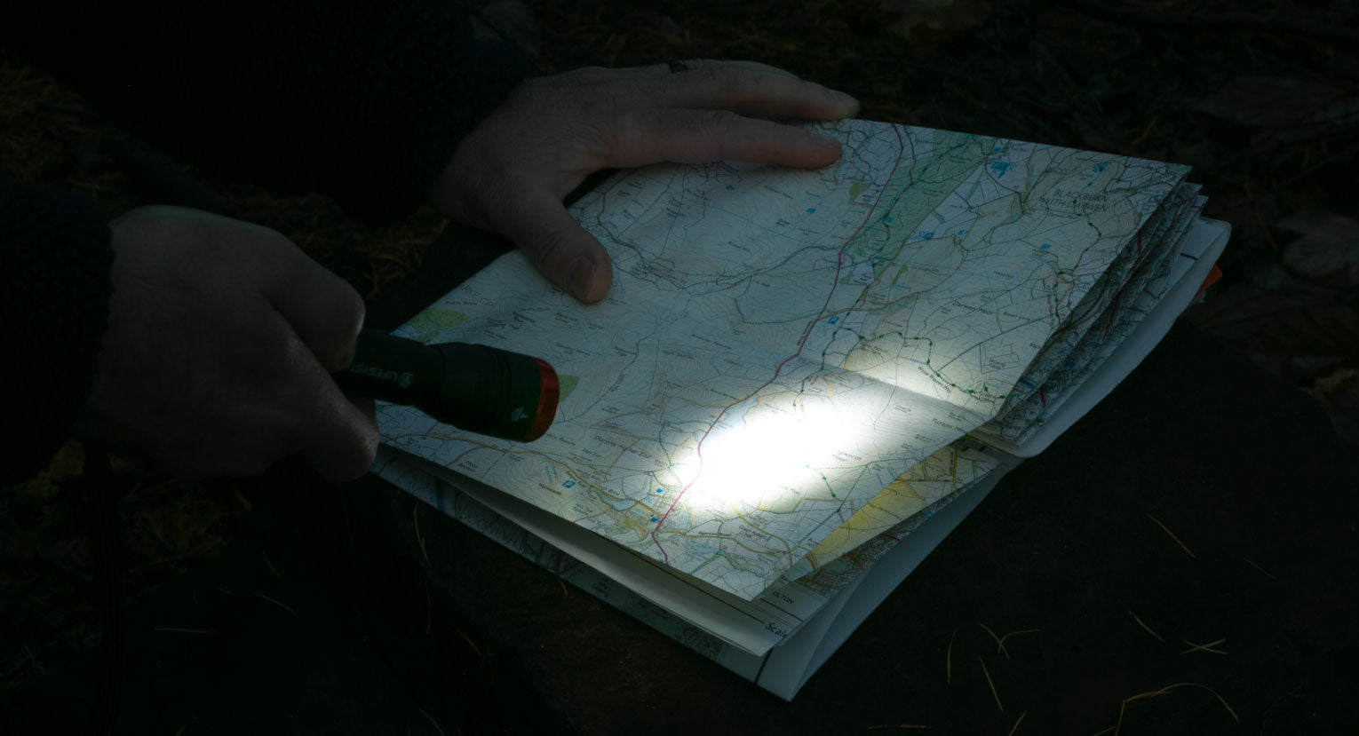 Person using a Lifesystems Intensity 545 hand torch to read a detailed topographic map in low-light conditions outdoors, with the torch beam illuminating a section of the map.